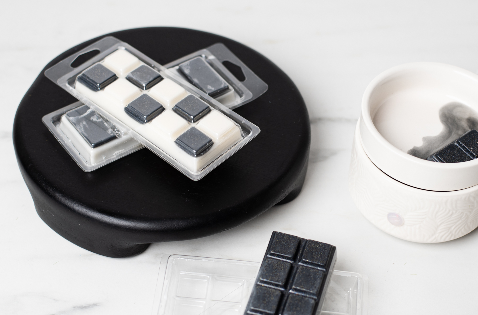 In the foreground, wax melt clamshells filled with black and white wax are stacked neatly. In the background, black wax melt squares are melting in a white wax melter.