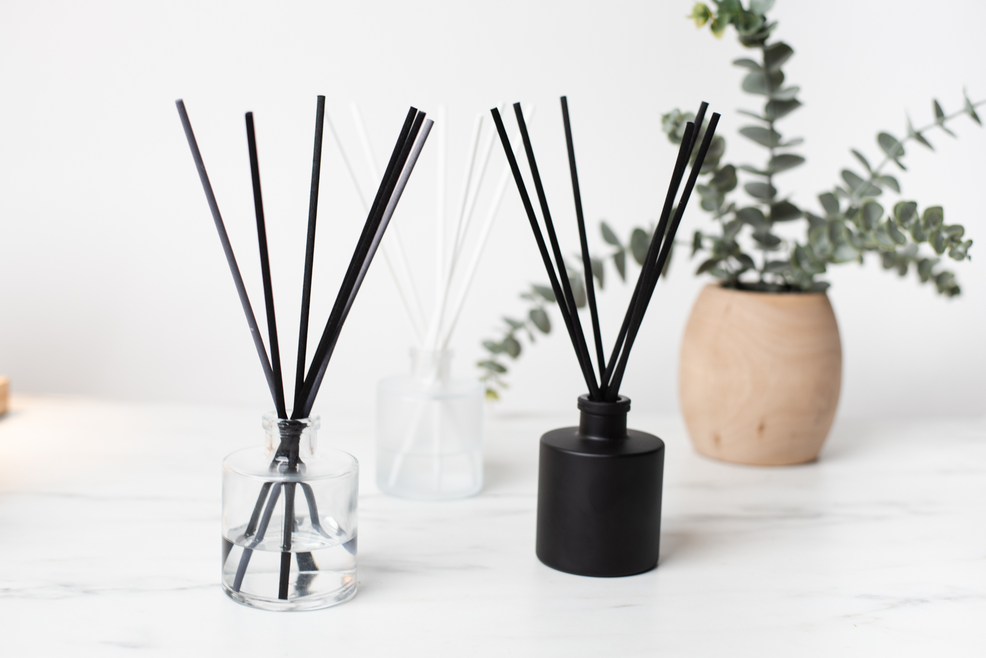 Three reed diffusers all in round jars, one is frosted glass, one is clear glass, and one is matte black. The reed diffusers are filled with black and white reed sticks. 