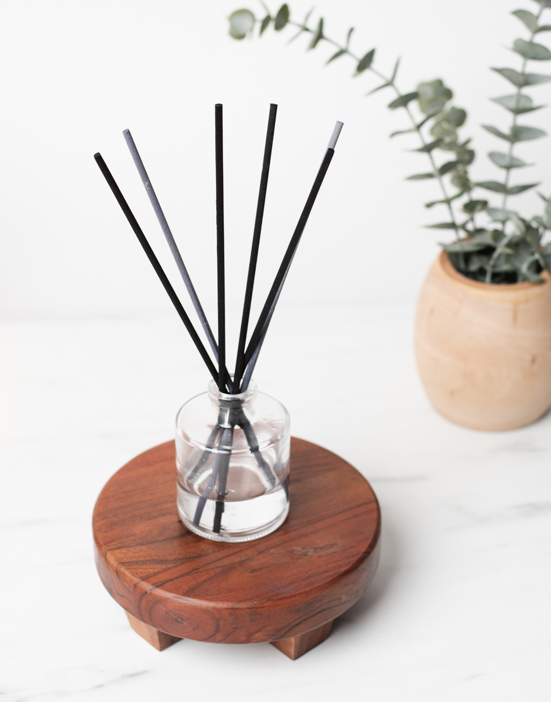 A reed diffuser in a clear round bottle rests atop a natural wooden stand. 