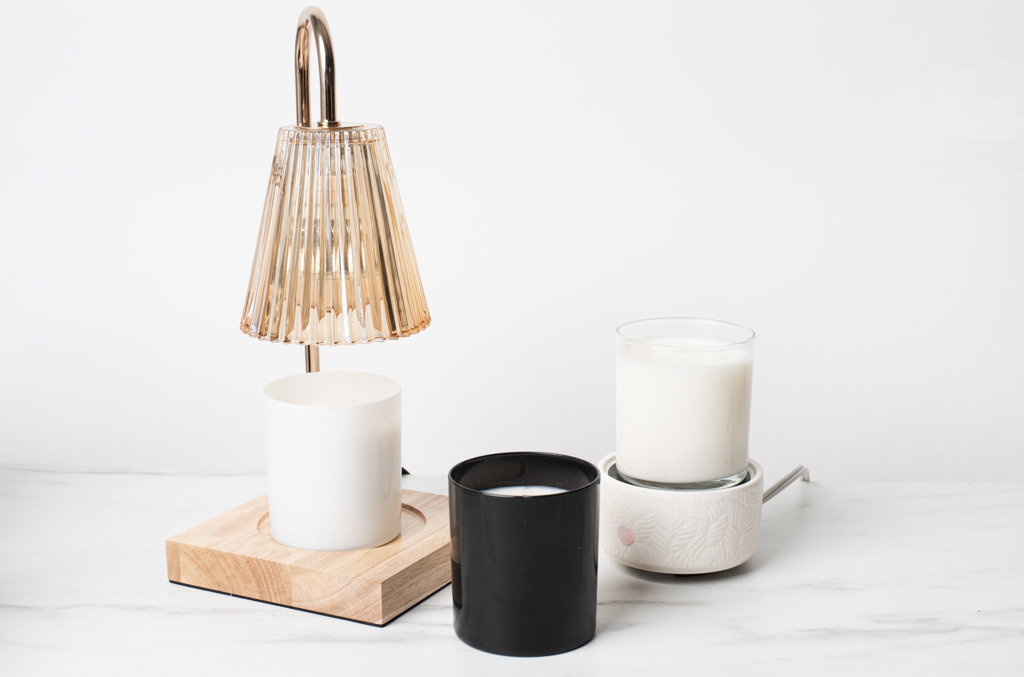 Three wickless candles, one white, one black, and one glass, on a marble surface. The white wickless candle is under a candle heating lamp.