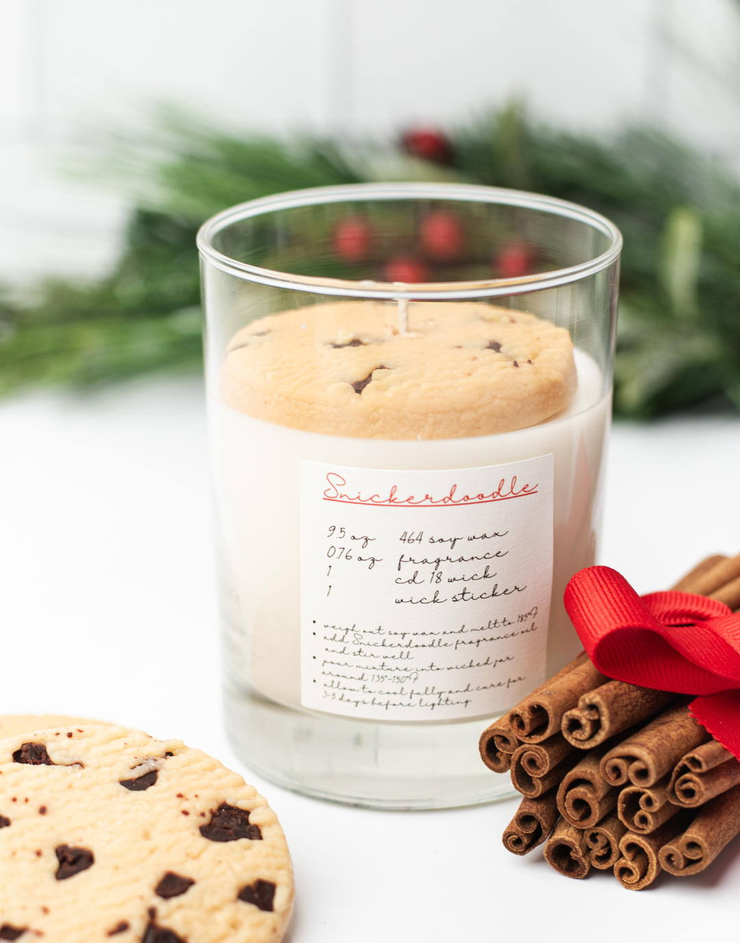 A candle that looks like a glass of milk with a cookie on top of it. On the candle, a white label reads "Snickerdoodle" and looks like a recipe. Beside the candle rests a bundle of cinnamon bark held together with red ribbon. 