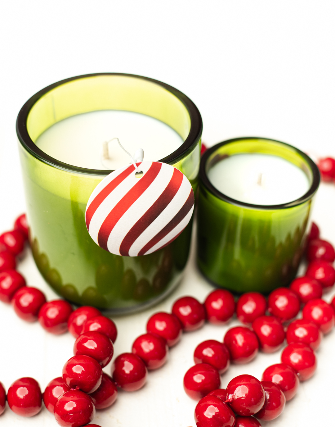 Two candles in green containers with red beads beside them. A tag that looks like a red ornament hangs from one of the candles.