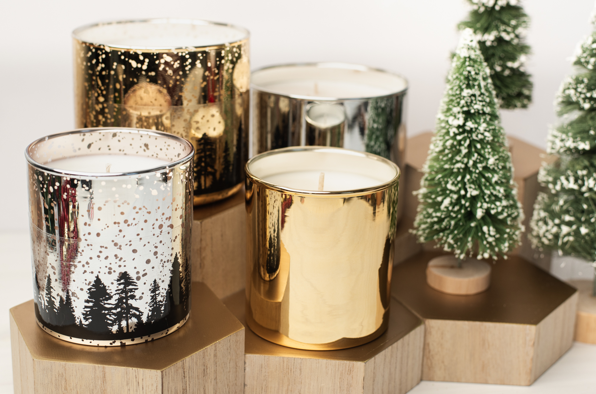 Candle containers in silver and gold beside small, snow-flocked Christmas tree figurines.