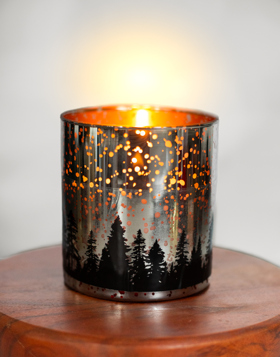 A silver three-wick candle with black tree-shaped images lining the bottom half of the container. The candle is lit and you can see light from the flame peeking through small areas of the container.