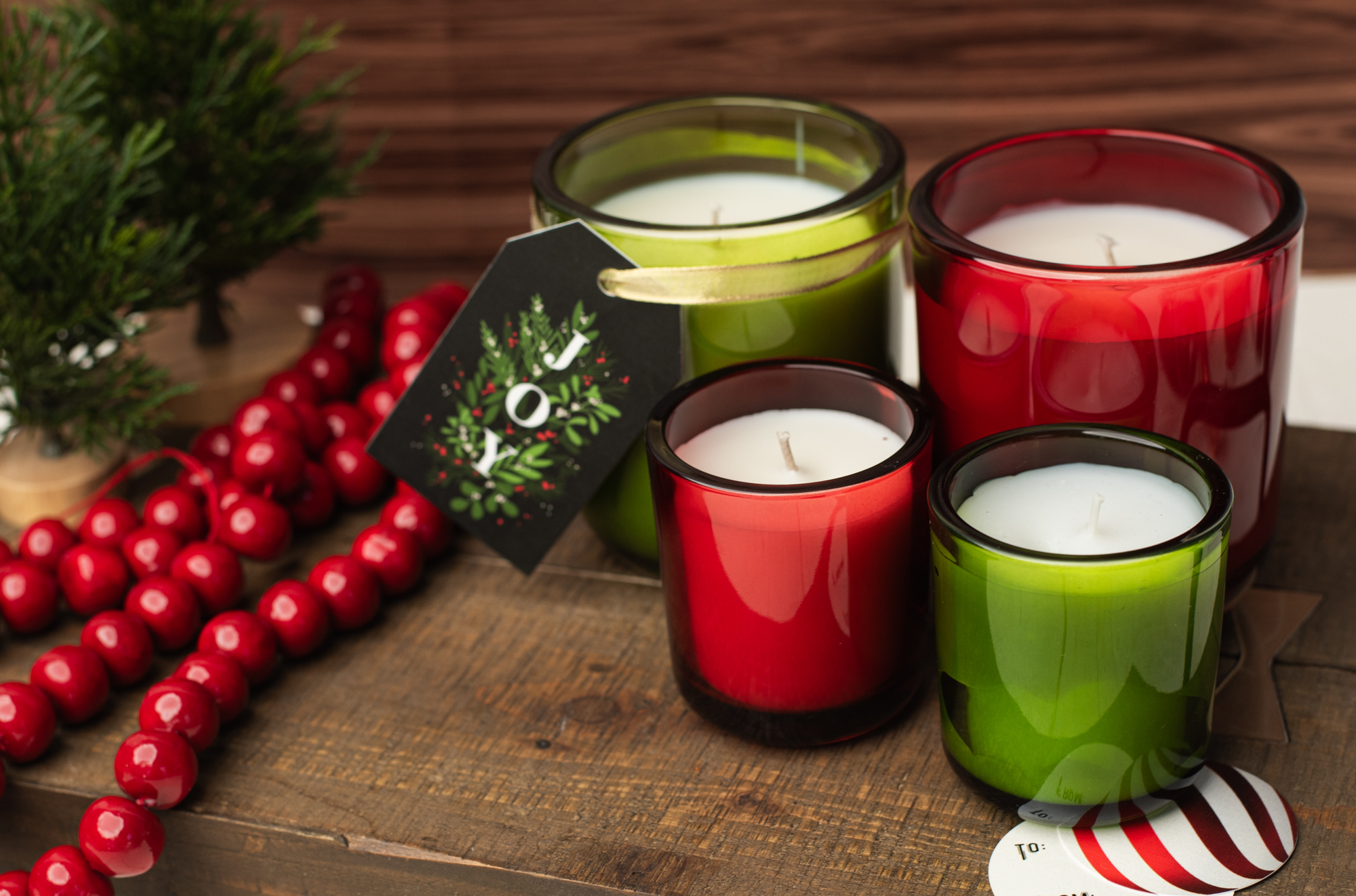 An assortment of candles in red and green containers. A tag that say "joy" in red and green hangs from one of the candles.