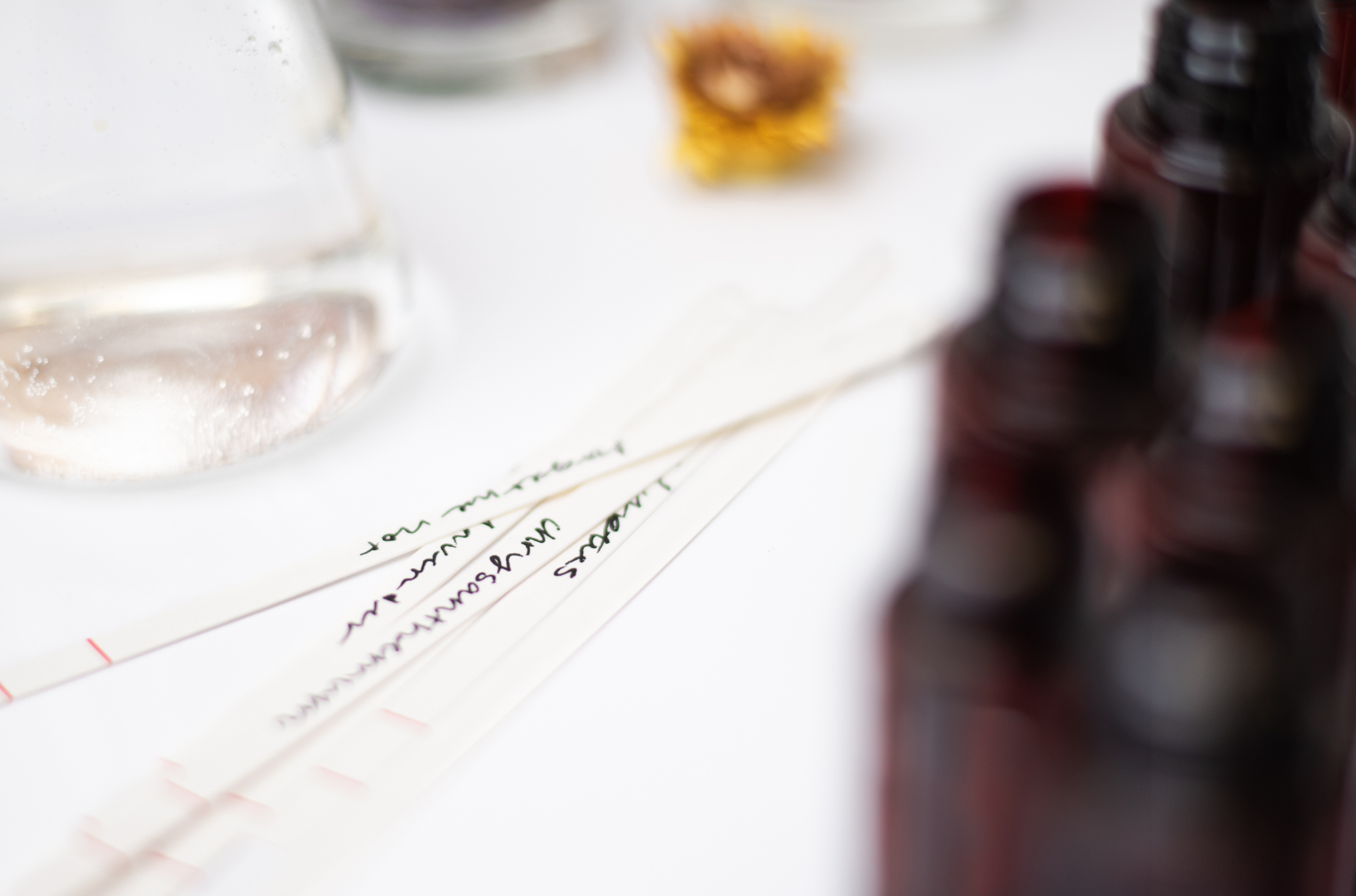Fragrance oil blotter strips are on a table beside various fragrance oils in amber bottles. 