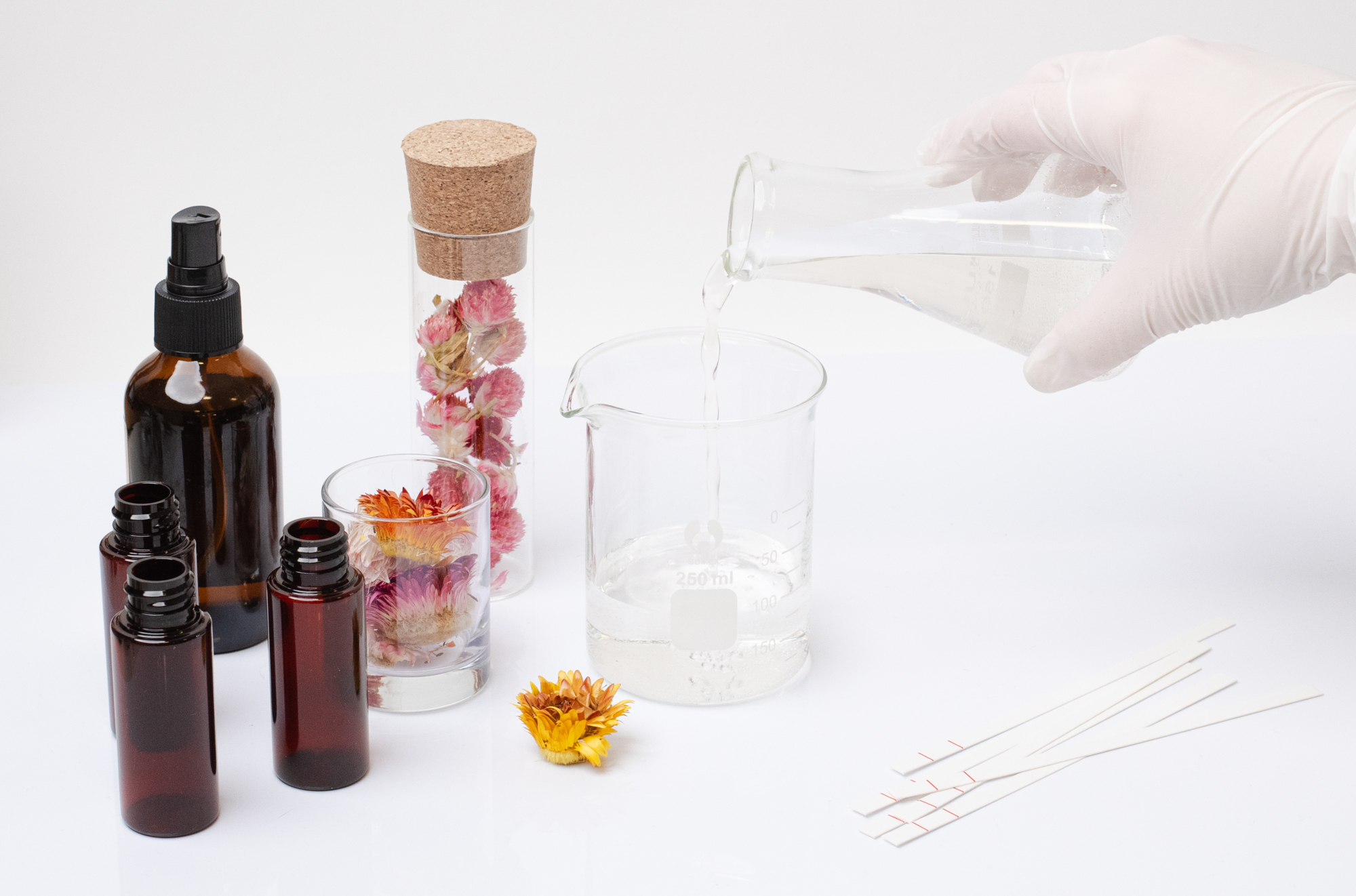 Someone is pouring fragrance oil into a small beaker. Beside the beaker sits a test tube filled with dried flowers and various luxury fragrance oil ingredients. 