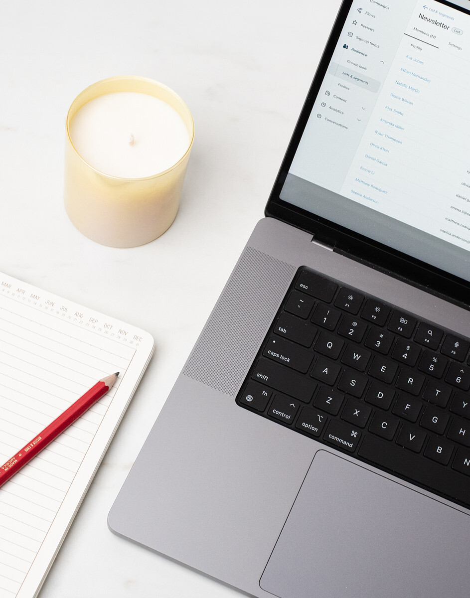 A silver laptop displays an email newsletter list; a notebook, pen, and candle sit next to the laptop