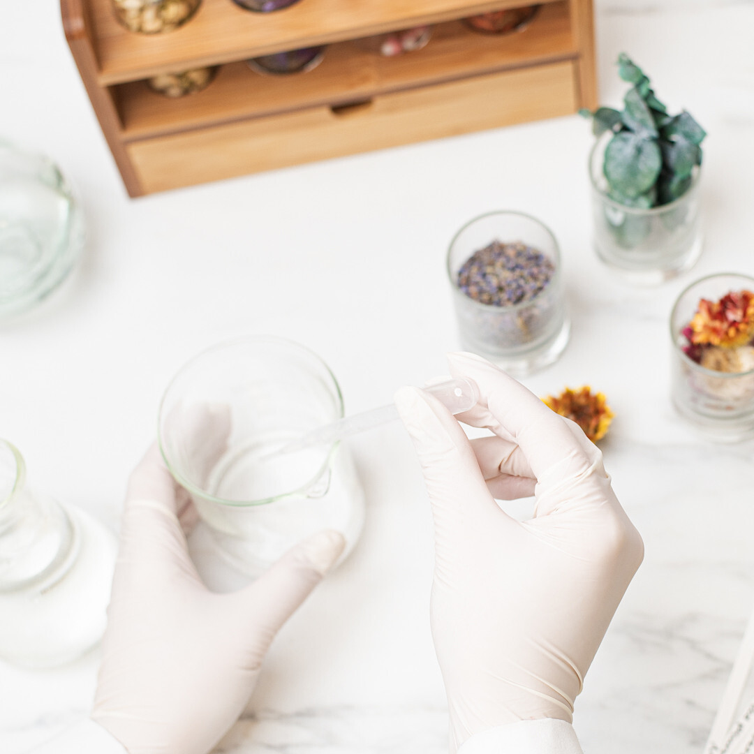 Someone wearing gloves is shown blending fragrance oils in a clear beaker. Amber fragrance oil bottles and other clear bottles containing clear liquid are also shown.
