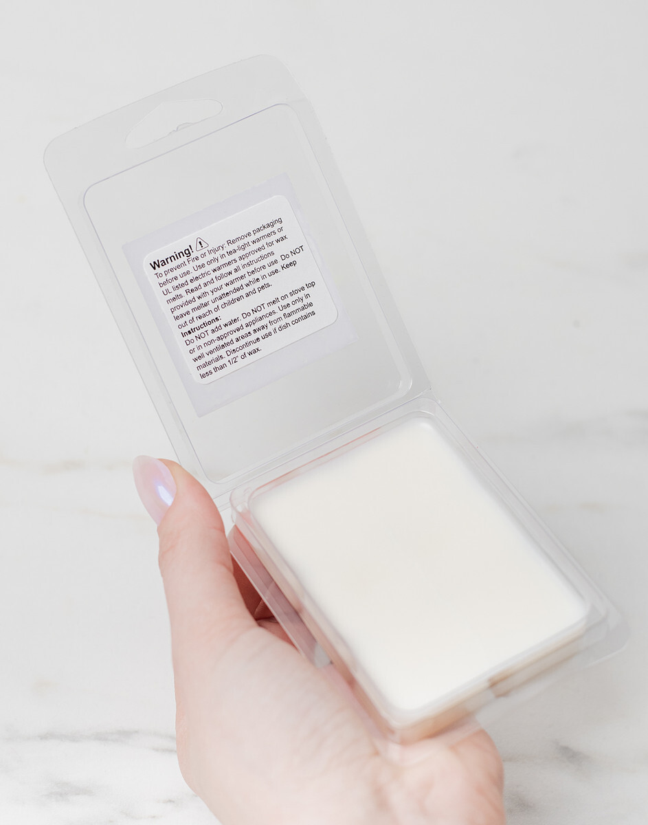 A light-skinned hand a Standard 6 pc Wax Melt Clamshell with its lid opened to reveal a Wax Melt Warning Label