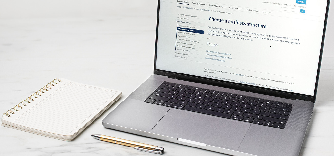 An open laptop displays a page from the Small Business Administration website title "Choose a business structure". Next to the laptop is a notebook and gold metal pen.