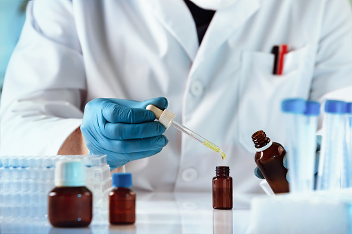 A close up view of a perfumer's hands wearing blue latex gloves as they use an eyedropper to place a golden liquid into a small amber glass bottle.