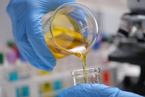 A close-up view of a pair of hands wearing blue latex gloves pouring a golden liquid from a beakers into a glass container.