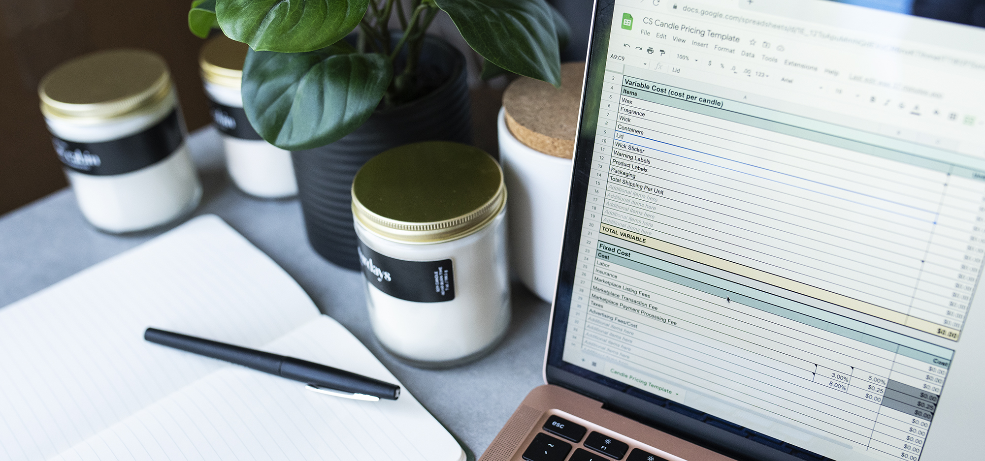 Candles, a notebook, and laptop displaying a candle pricing spreadsheet