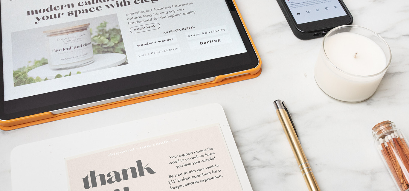 On a marble surface an iPad displays a candle brand website's home page and an "as featured in" section, a thank you card order insert, a cell phone showing an Instagram post with a customer review, a candle in a glass jar, a golden metal pen, and a glass bottle of matches.