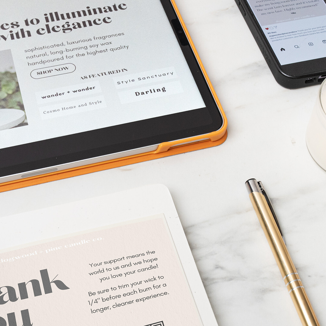 On a marble surface an iPad displays a candle brand website's home page and an "as featured in" section, a thank you card order insert, a cell phone showing an Instagram post with a customer review, a candle in a glass jar, a golden metal pen, and a glass bottle of matches.