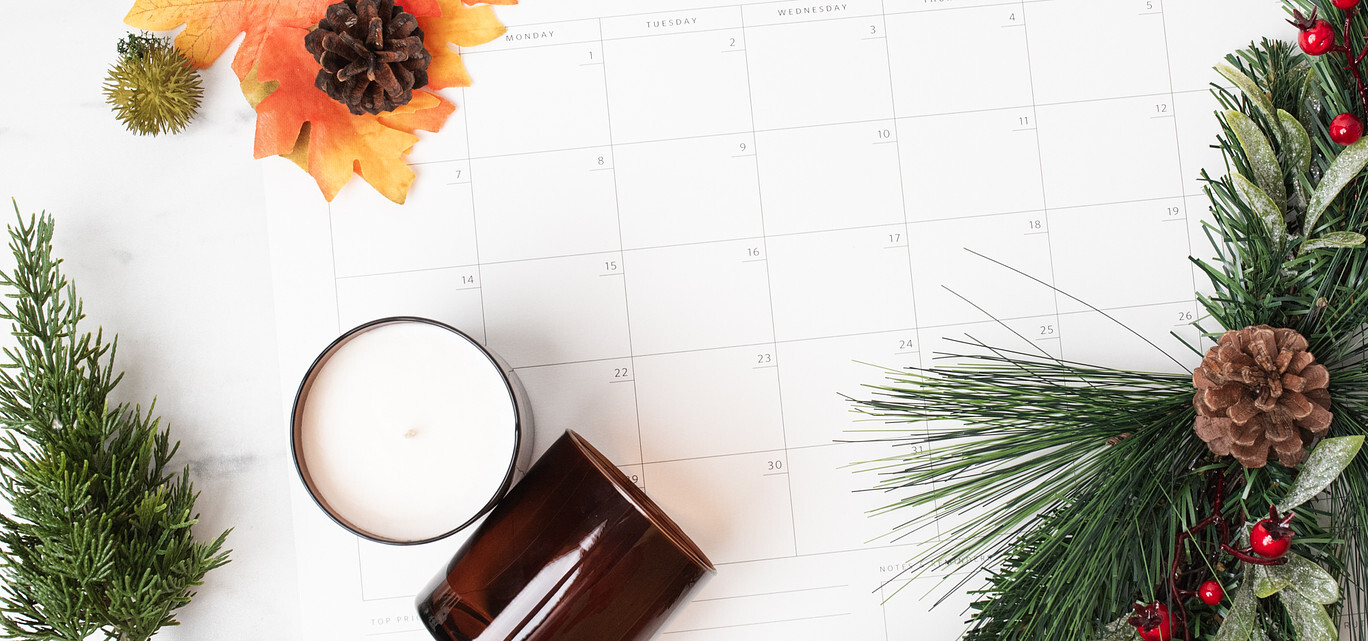 Winter greenery, fall foliage, and two candles in amber glass tumbler jars frame a desktop paper calendar displaying a month view. 