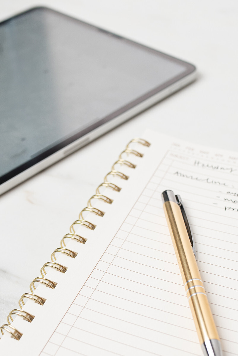 A gold metal pen sits atop a notebook next to an iPad.