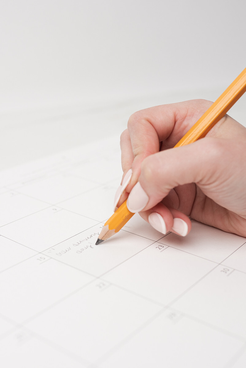 A light-skinned hand writes a note onto a paper calendar with a pencil.