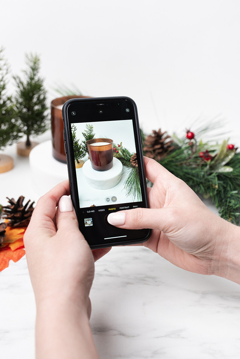 A pair of light-skinned takes a photo of handmade candle in a holiday vignette on a mobile phone.