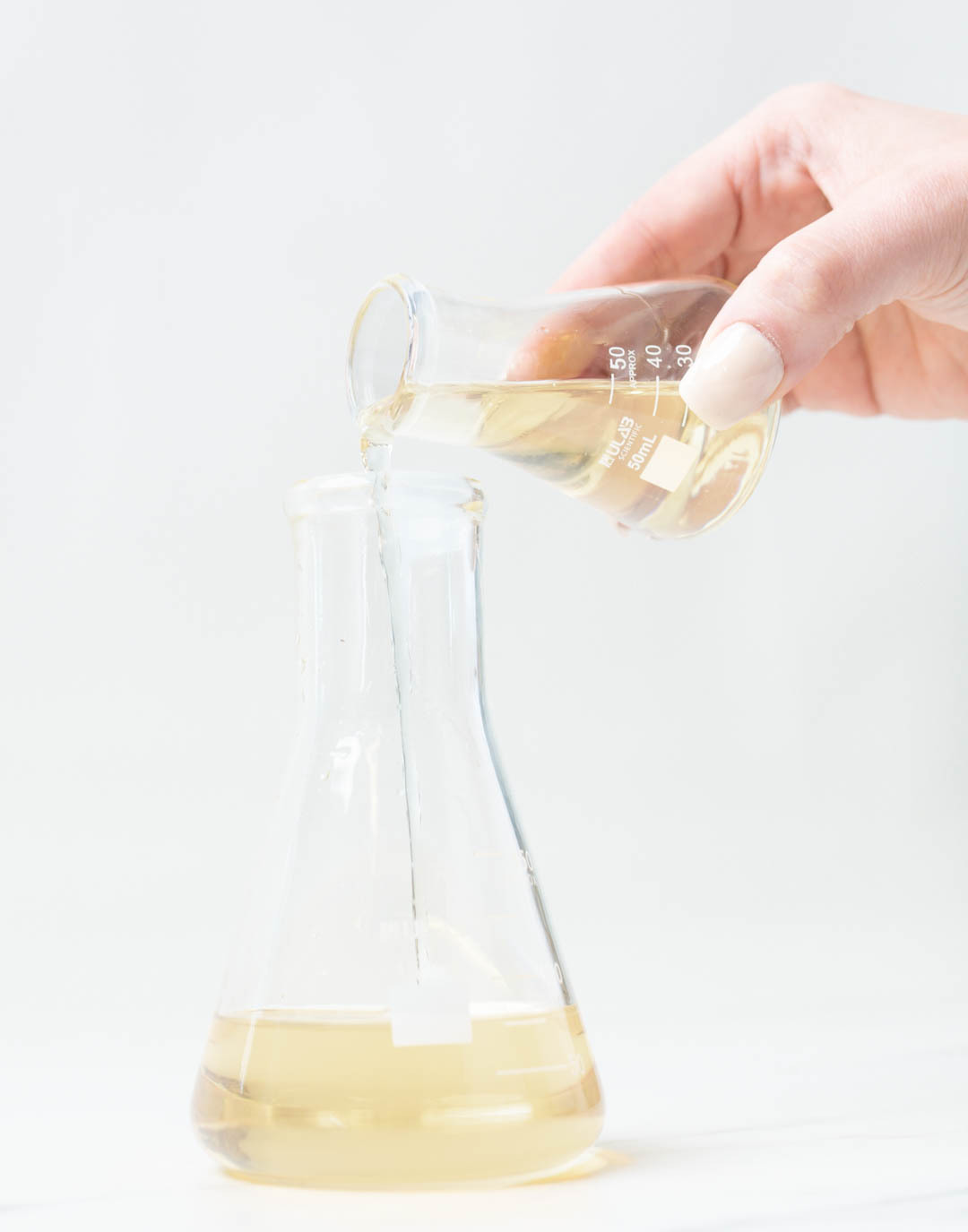 Someone is shown pouring fragrance oil from one laboratory glass flanks into another. 