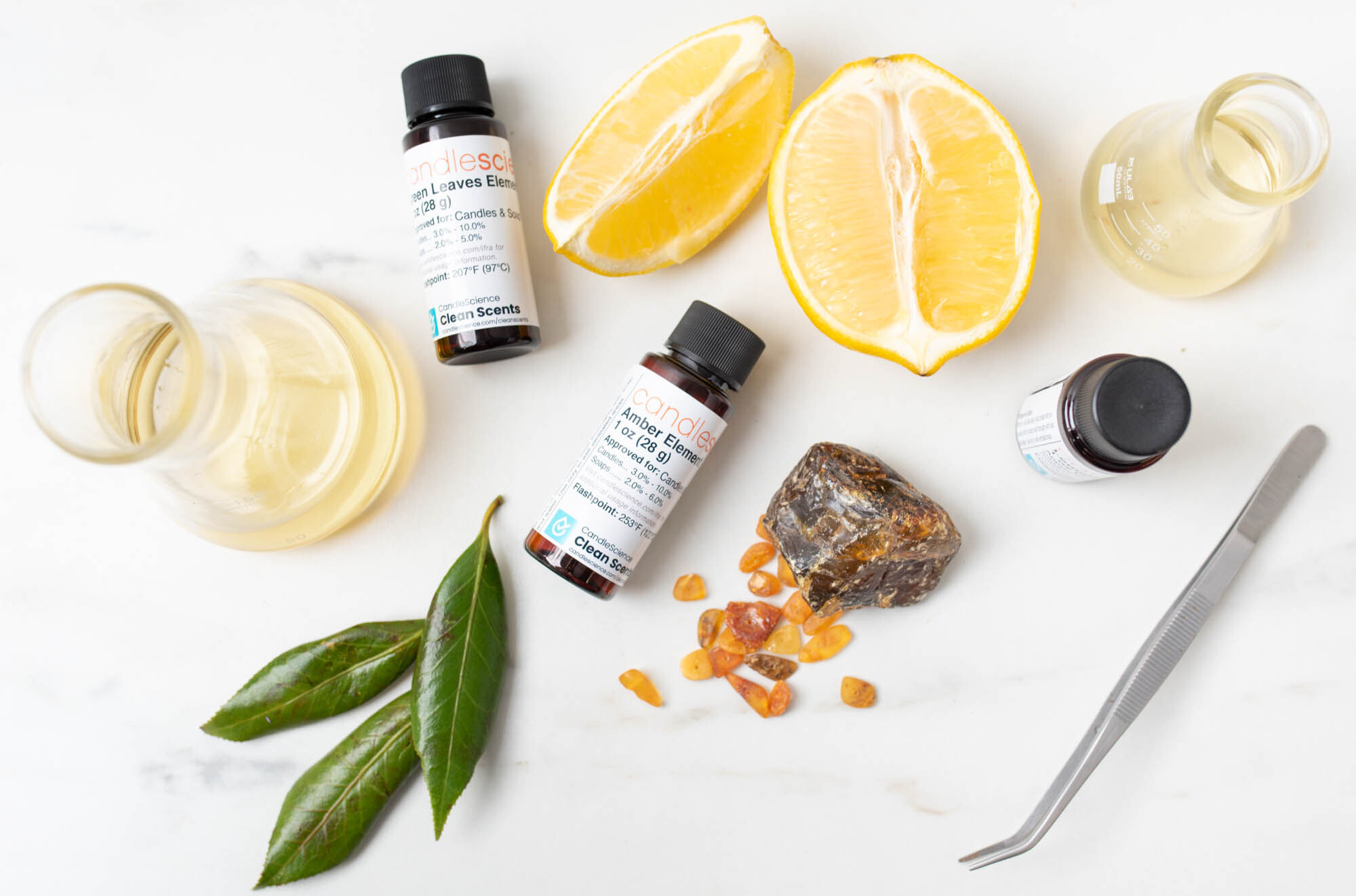 A flat lay of fragrance oil blending ingredients on a marble surface. The items include two small amber-colored fragrance oil bottles, a sliced lemon, a piece of amber resin with small chips beside it, green leaves, two laboratory glass flasks containing liquid, and a pair of metal tweezers.