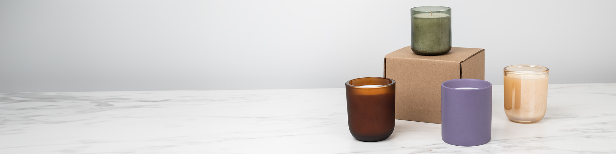 Three candle containers in green, frosted amber, and purple on a white marble countertop with a single piece shipping box in the backdrop.