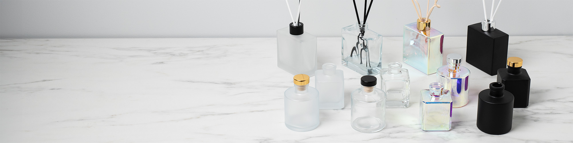 A variety of frosted, prism, and matte black reed diffuser bottles with reeds on display on white marble countertop.