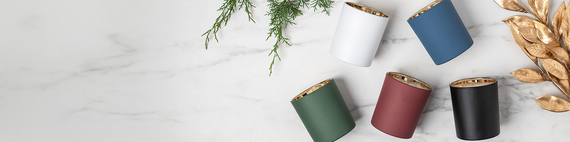 white, blue, red, green, and black tumbler jars with a warm gold interior displayed on a white marble countertop with evergreen and gold leaves. 