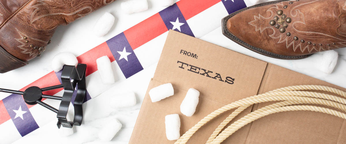 Texas-themed arrangement featuring a shipping box stamped "from: Texas." It includes brown cowboy boots with decorative stitching, a lasso rope, and packing peanuts. There is also a strip of a Texas flag design featuring stars, and a branding iron tool visible to the side. The overall theme emphasizes Texas culture and western motifs.