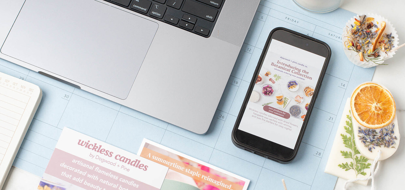 An overhead view of a desk with a cell phone displaying a wholesale email on its screen, a laptop, Dogwood + Pine Candle Co. shelf talkers, and assorted wax products.