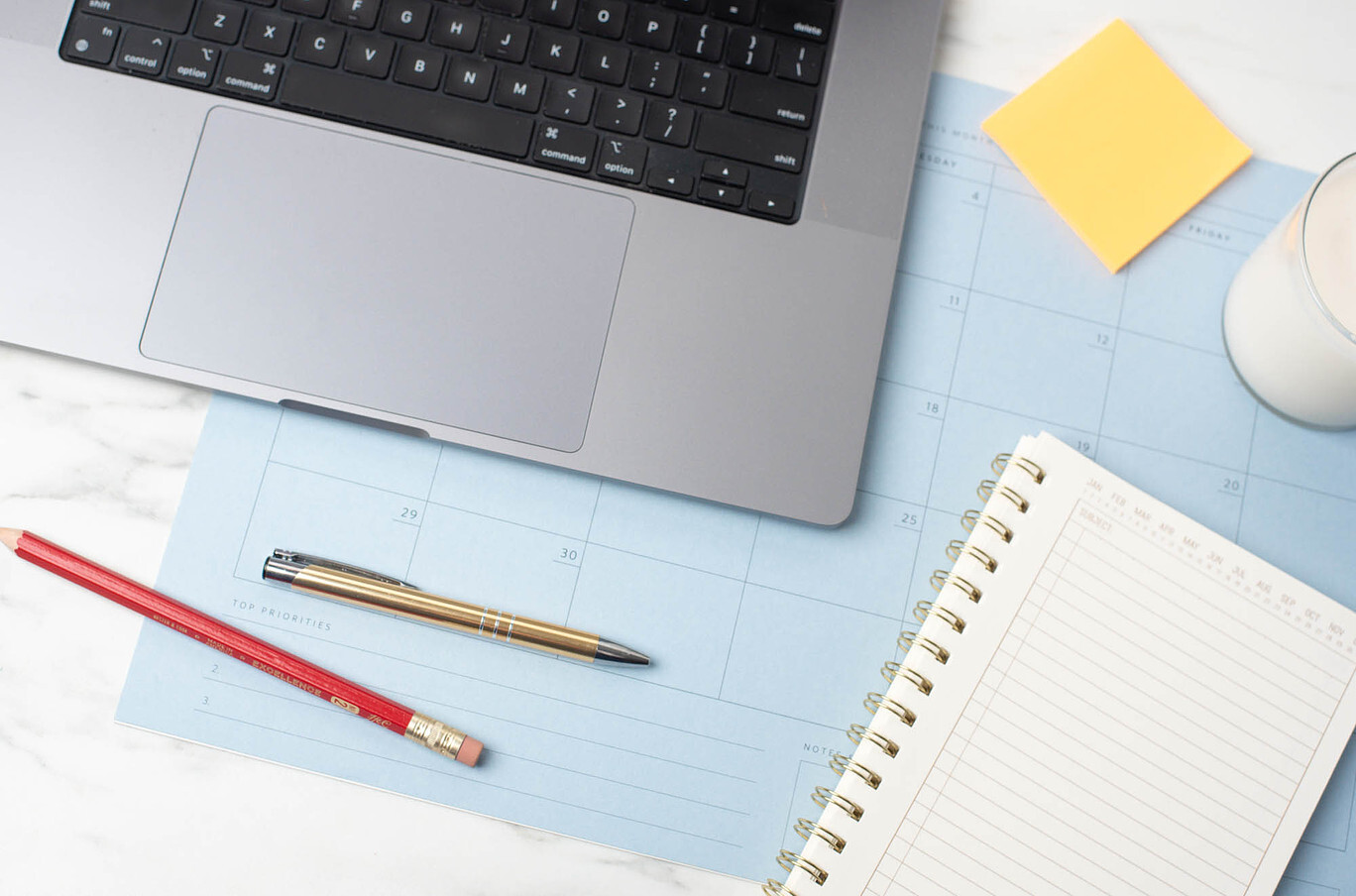 A silver laptop, notebook, pens, and a candle sit on a desktop