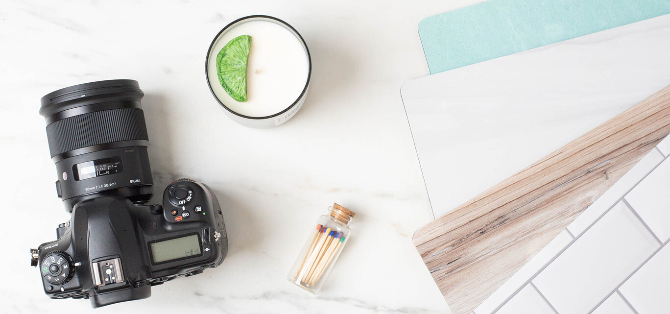 A DSLR camera lays on a tabletop next to a handmade candle, a small jar of matches, a fanned out selection of photography surfaces.