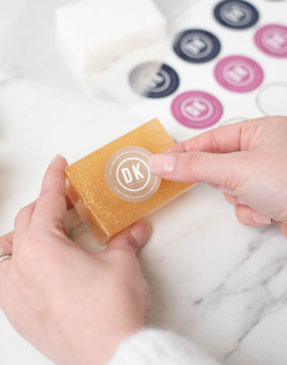 A pair of light-skinned hands places a round label on a bar of melt and pour soap.