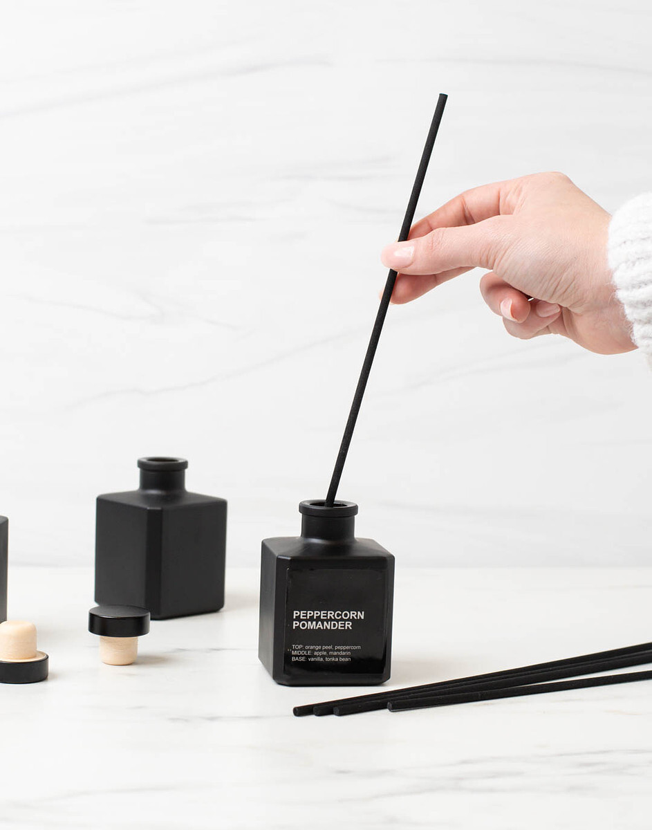 A light-skinned hand places a black reed diffuser stick into a matte black reed diffuser bottle.