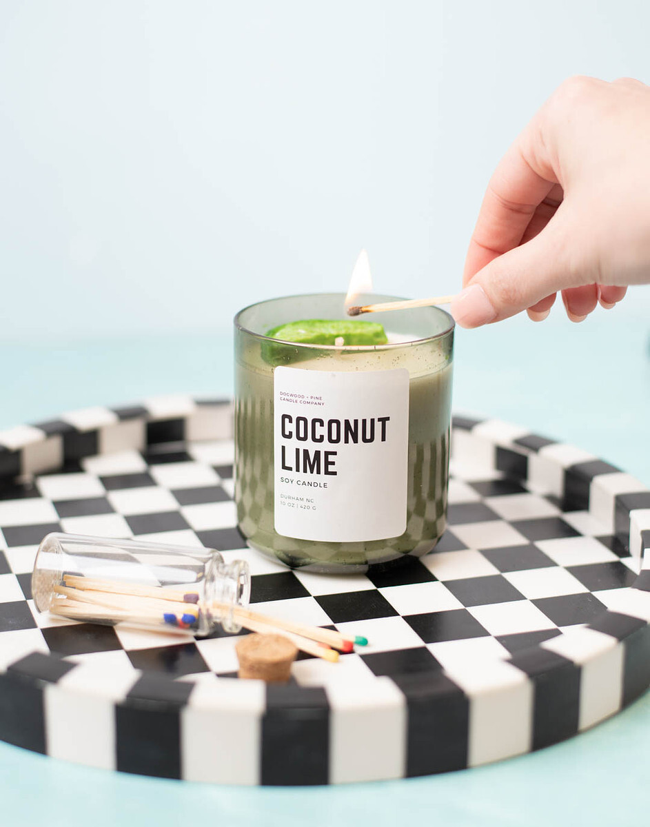 A light-skinned hand holding a lit match to the wick of a candle in a Smoked Olive Bubble Tumbler Jar.