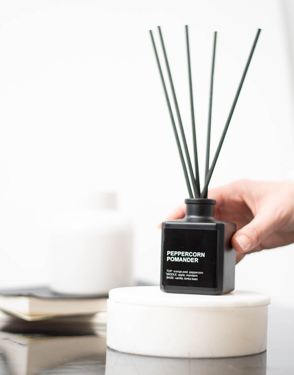 A light-skinned hand holds a matte black reed diffuser bottle filled with with black reed diffuser sticks.