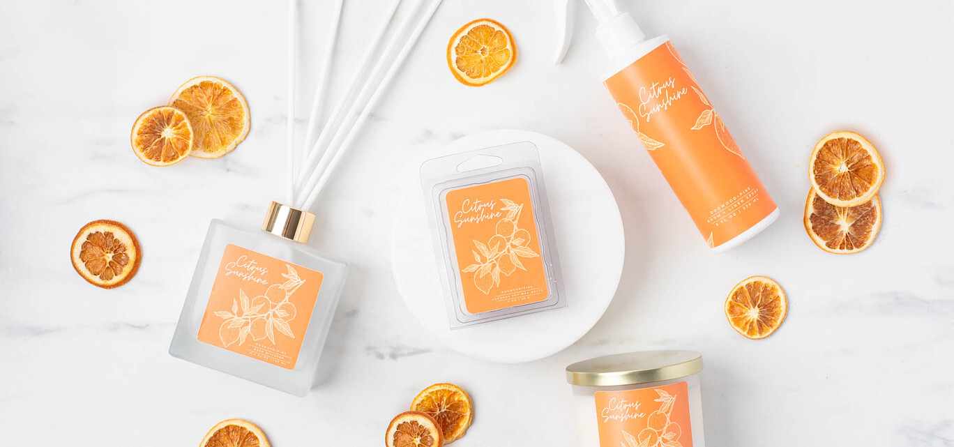 A flatlay view of a frosted reed diffuser bottle with white reed diffuser sticks, a clear wax melt clamshell, and a white spray bottle of room and linen spray, all featuring orange labels. 