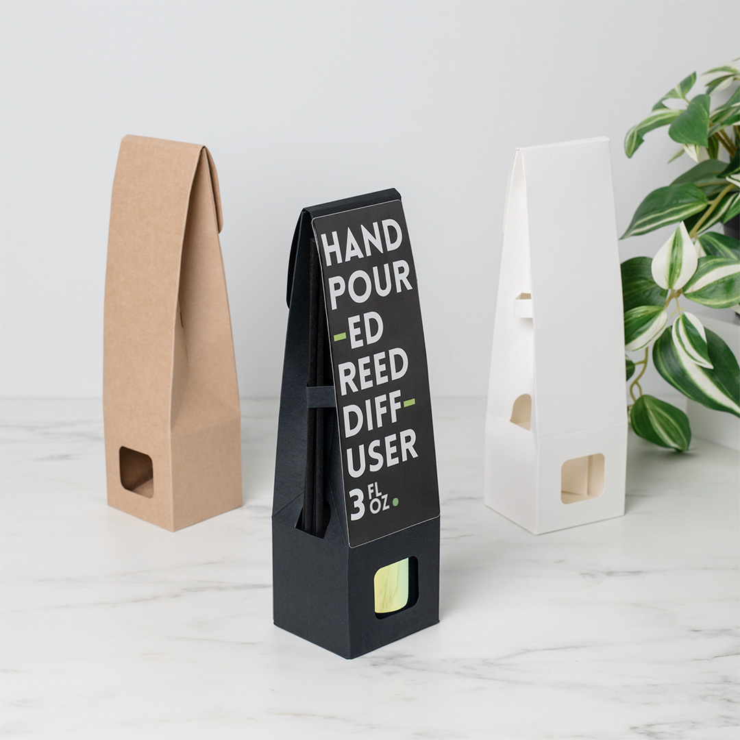 A labeled black retail reed diffuser packaging for a round reed diffuser bottle sitting between two unlabeled kraft and white boxes. 