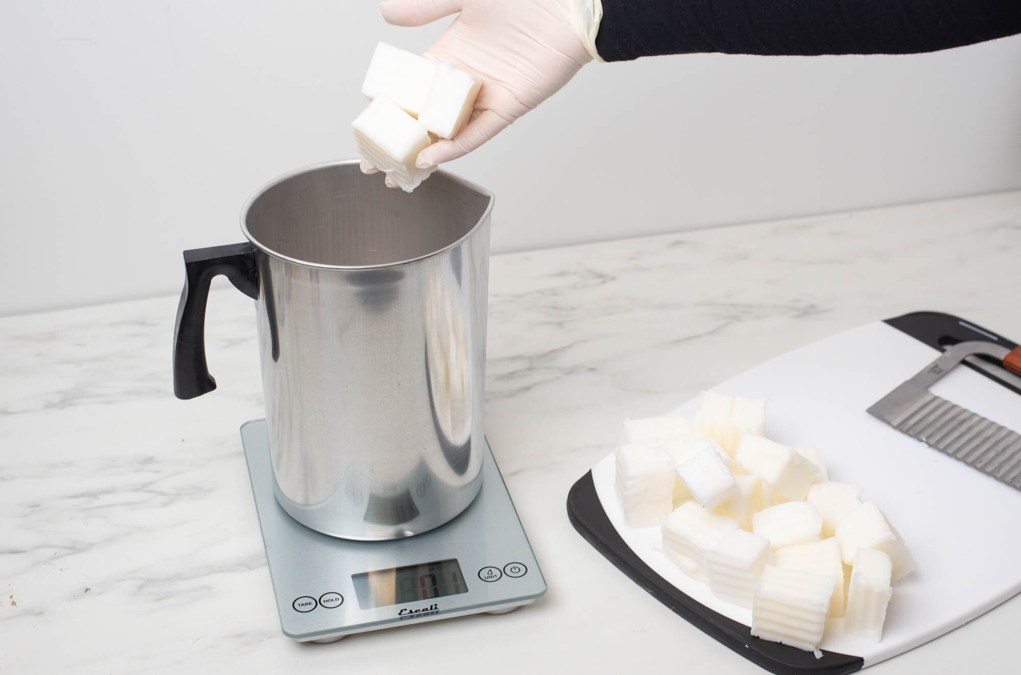 Cubed wax being placed into a pouring pitcher on a scale next to a cutting board.