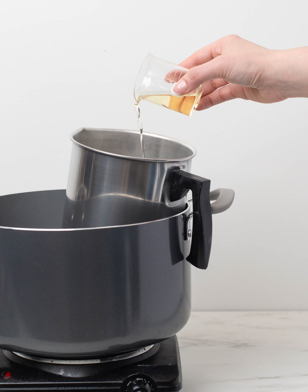 A hand pouring fragrance oil from a glass measuring cup into a pouring pitcher in a double boiler.