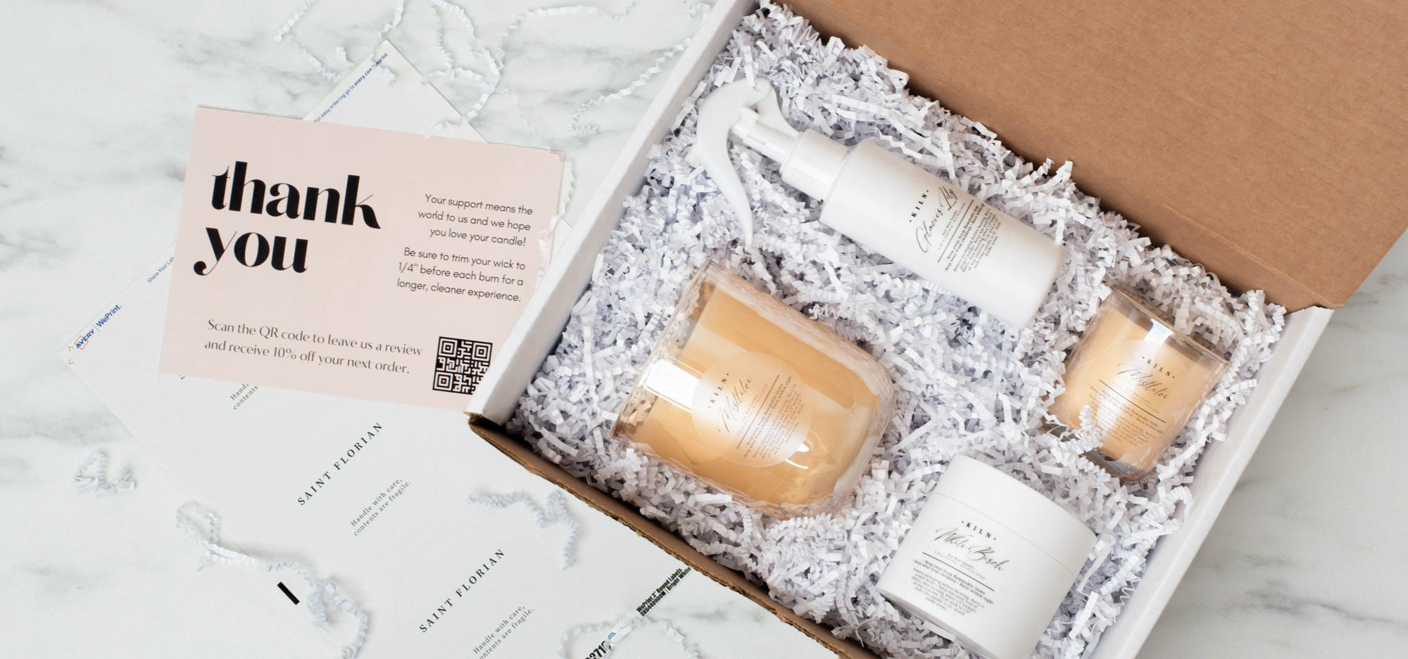 Two candles, a bottle of room spray, and a jar of sugar scrub nestled in an open box filled with white crinkle paper, and a thank you note next to the box 