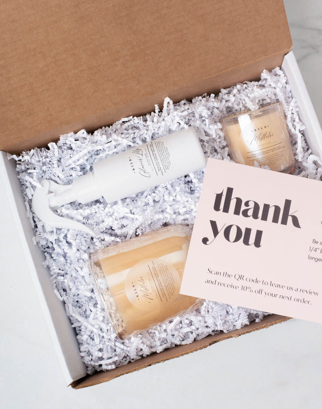 Two candles, a bottle of room spray, and a jar of sugar scrub nestled in an open box filled with white crinkle paper, and a thank you note on top of the products