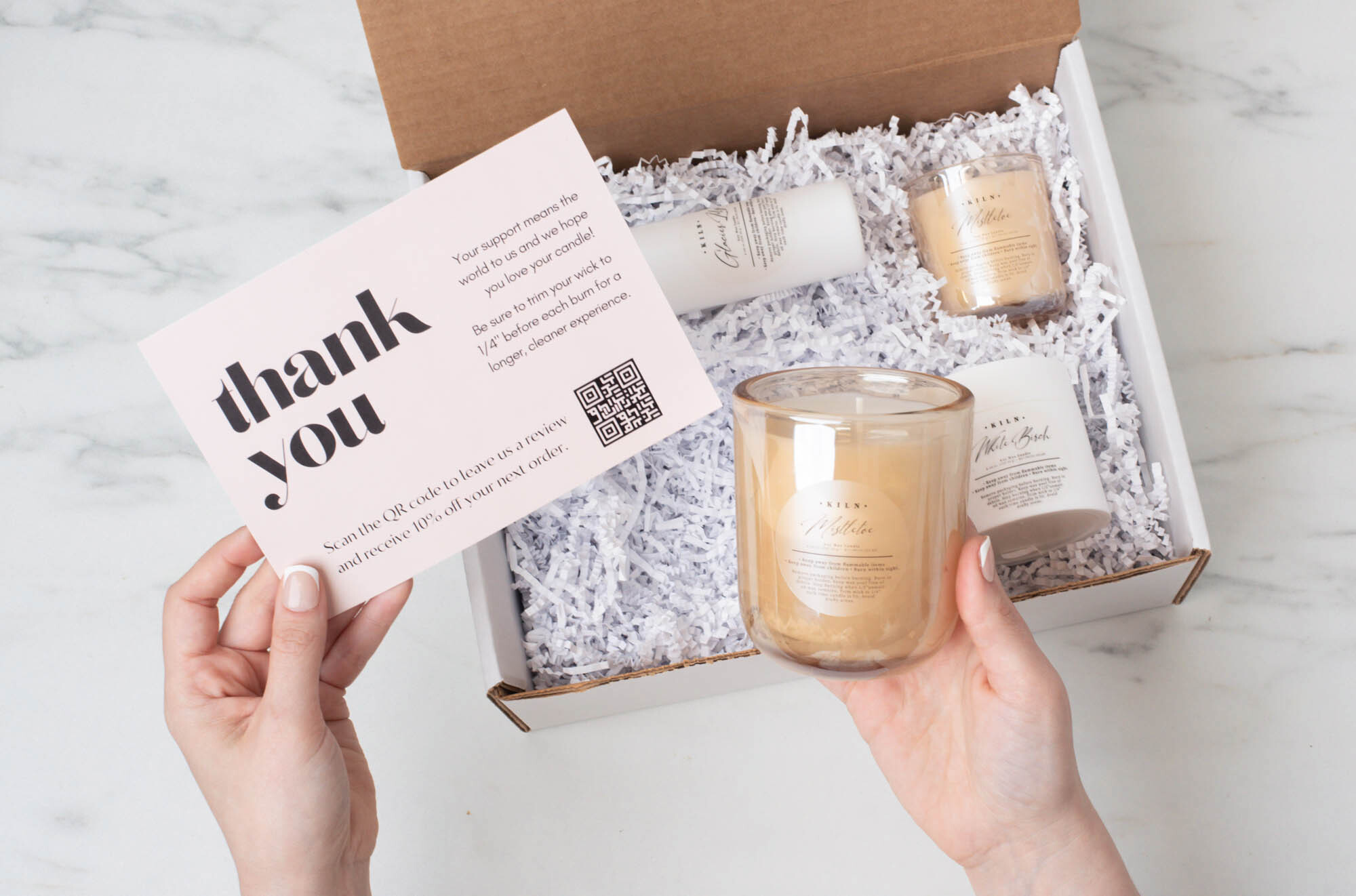 A pair of light-skinned hands hold a thank note and a candle jar above an open box filled with white crinkle paper