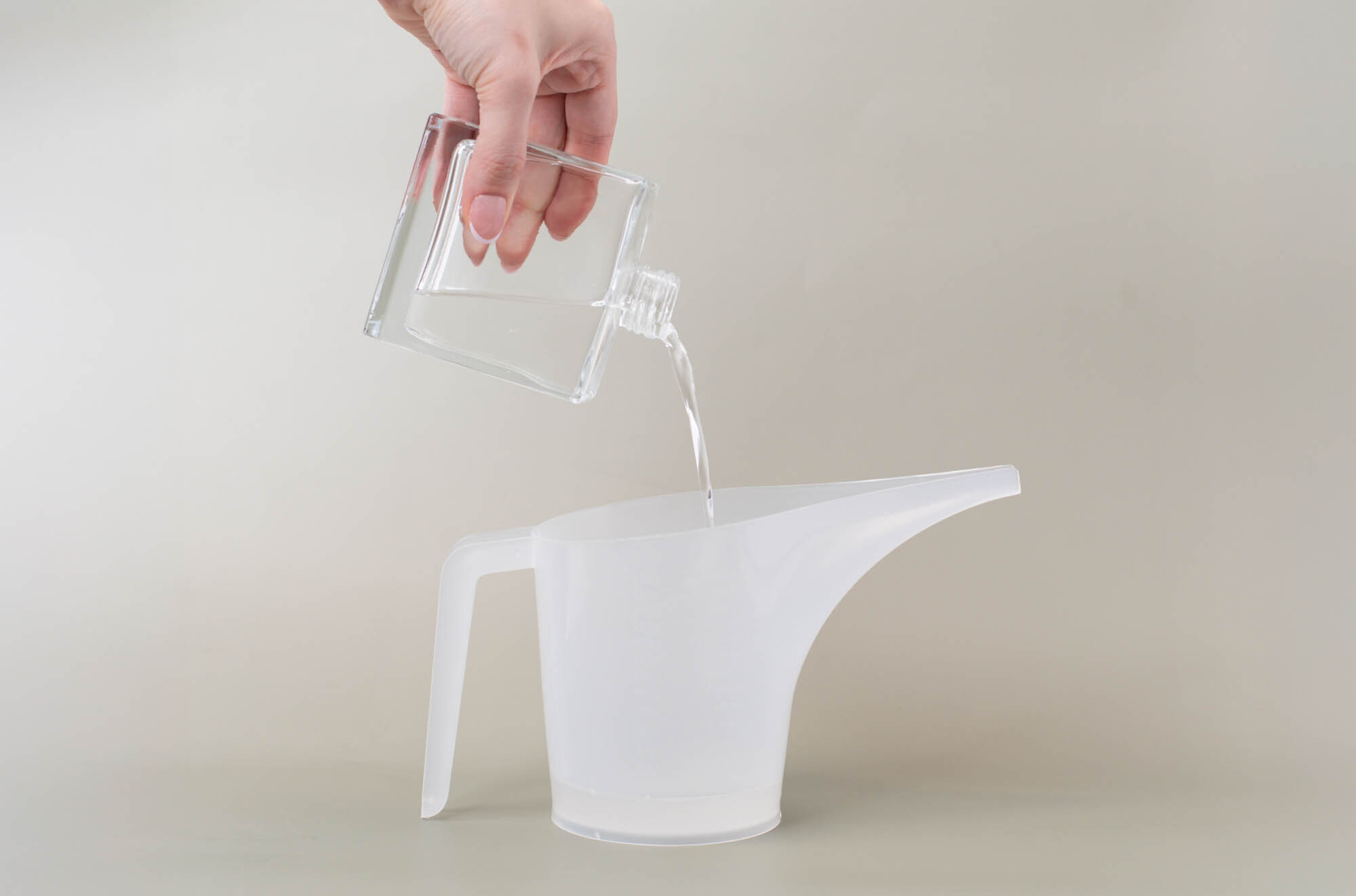 A hand pouring liquid out of a glass reed diffuser into a funnel pouring pitcher