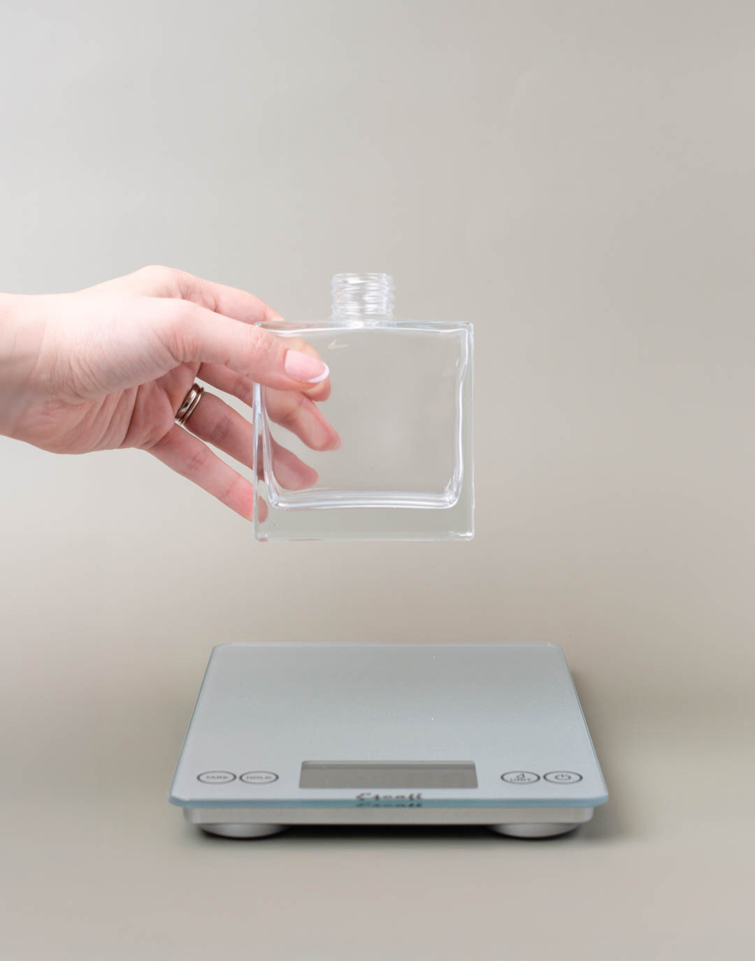A hand placing an empty glass reed diffuser onto a scale.