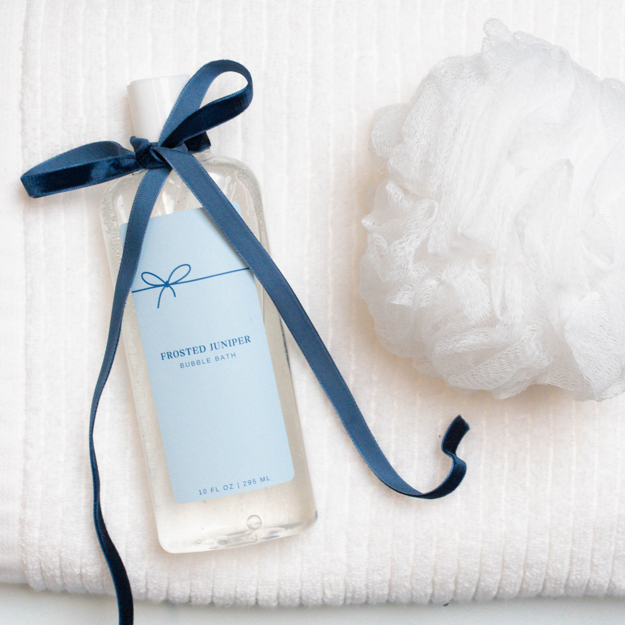Bottle of bubble bath with a ribbon on a white towel next to a white loofah. 