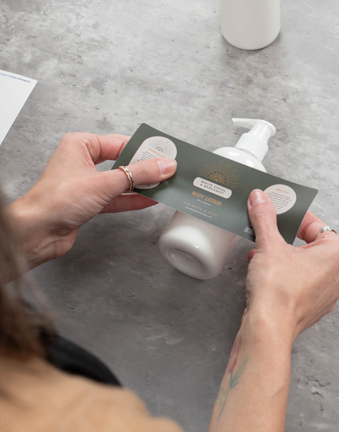 A pair of light-skinned hands applies a product label to a bottle of lotion made with CandleScience supplies