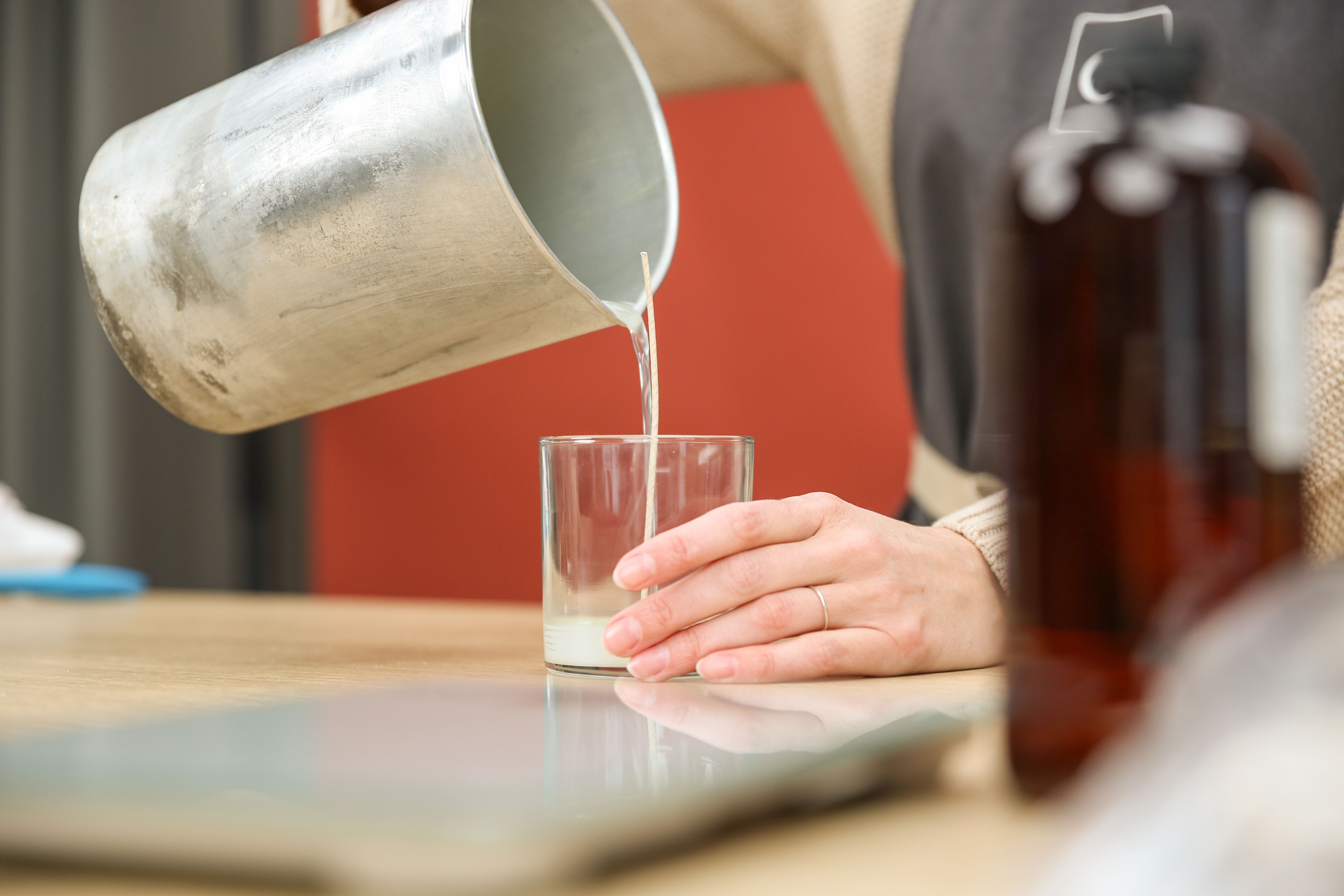 A pair of light-skinned hands holds a metal pouring pitcher and dispenses melted wax into a glass candle jar