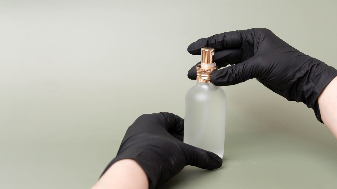 Gloved hands screwing a pump onto a frosted perfume bottle.